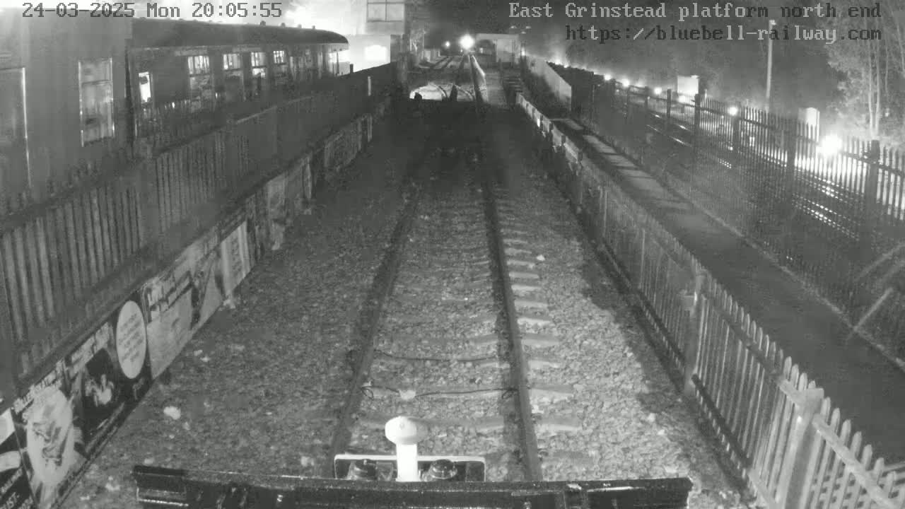 A nighttime view of a railway platform with a train car partially visible, railway tracks extending into the distance, and fences lining the platform edges.