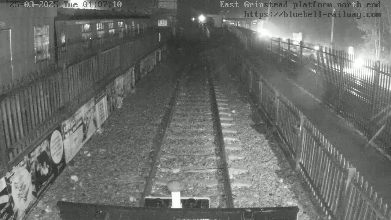 A nighttime, low-light view of a train platform with railway tracks extending into the distance, bordered by fences and posters.