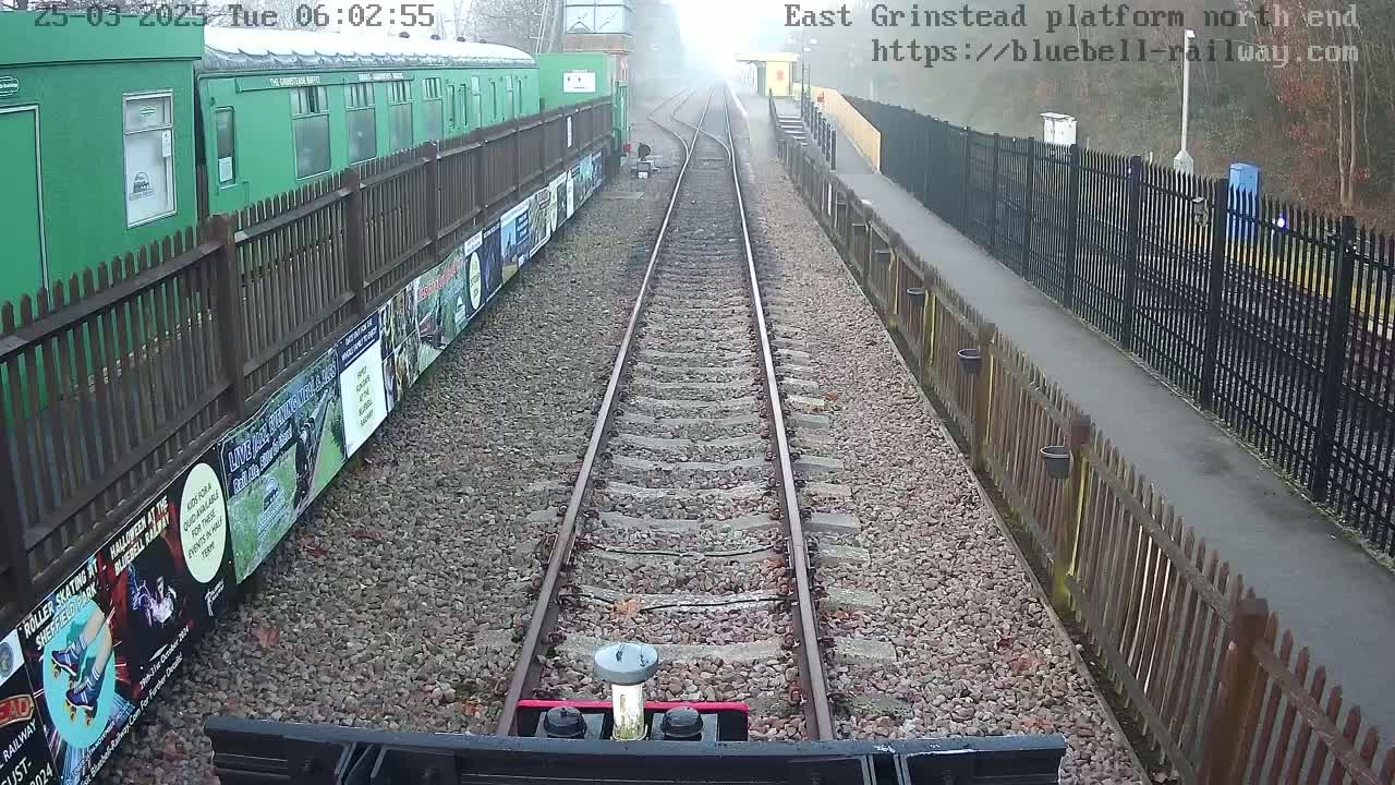 A view from a train on a railway track on an overcast day, alongside a platform with a green train car and wooden fences.