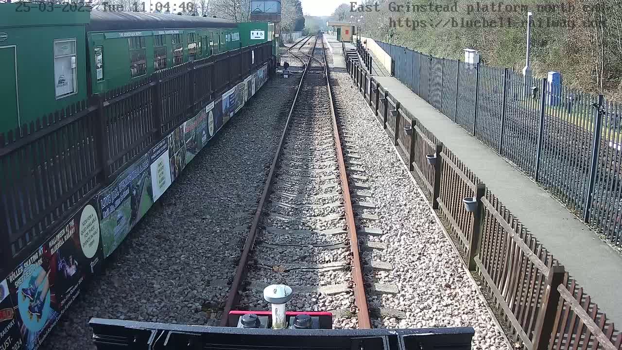 A view down a railway track on a sunny day, with a green train car and posters on the side visible to the left.