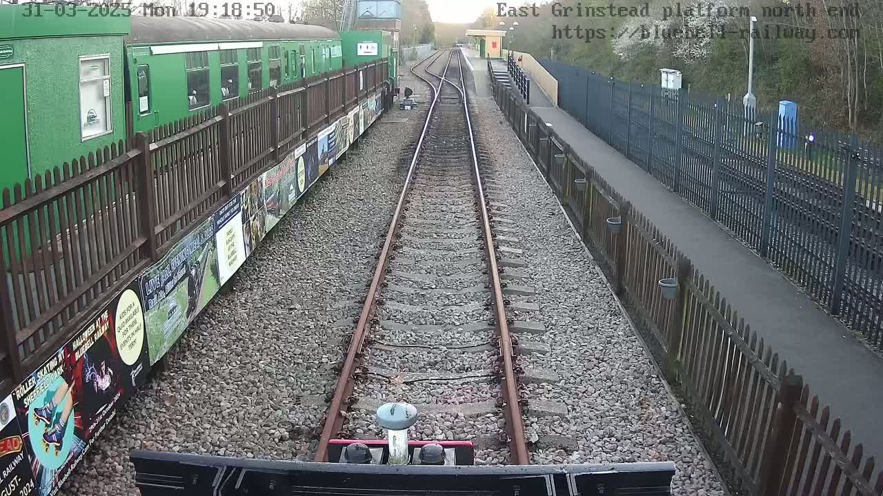 A train is approaching a railway station platform on a clear evening, with a green train car visible to the side.