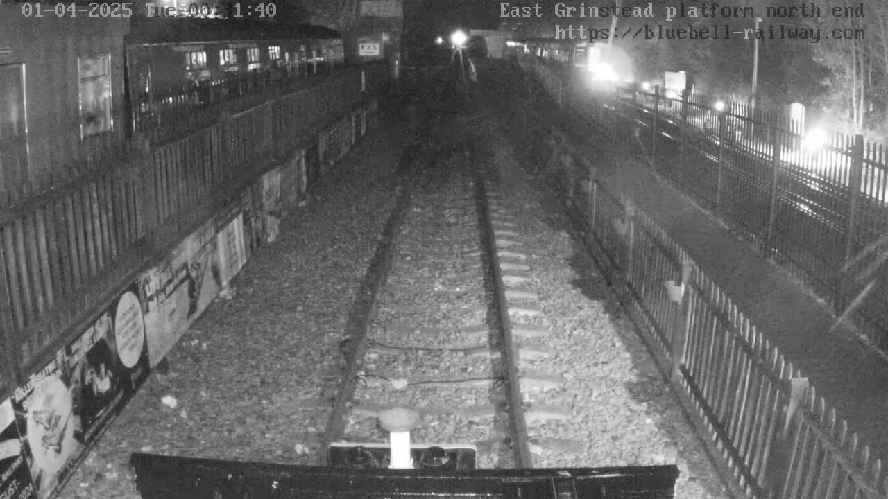 A nighttime view of train tracks at a platform, bordered by fences and posters, under dark conditions.