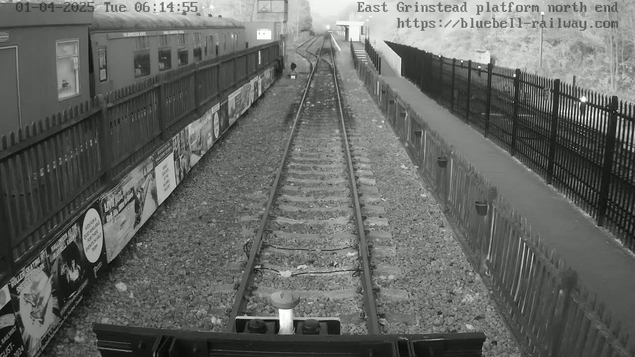 A grayscale view from a train shows a railway platform with a train alongside, bordered by fences and advertisements, on a cloudy day.