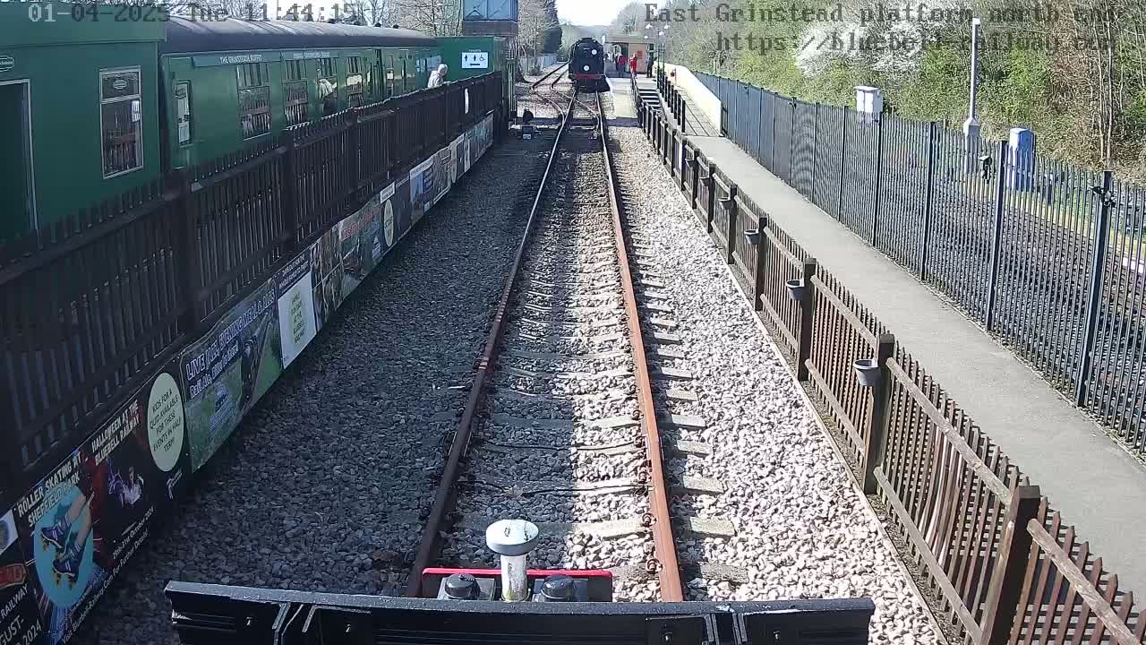 A dark-green train is approaching a station on a sunny day, viewed from the perspective of a vehicle on the tracks.