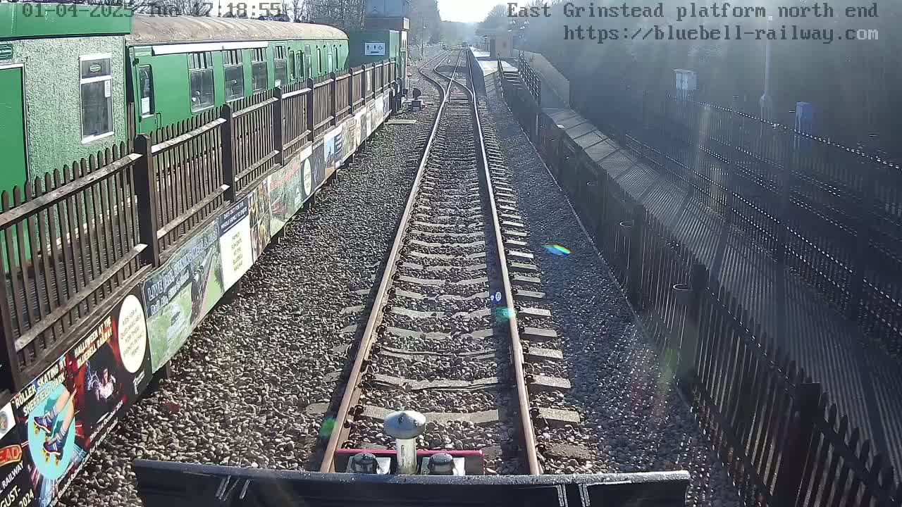 A view from a train down a railway track on a sunny day, with a green train and wooden fences on either side.