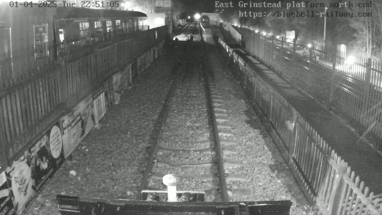 A nighttime view from a train shows railway tracks extending into a tunnel, flanked by fences and platforms with posters.