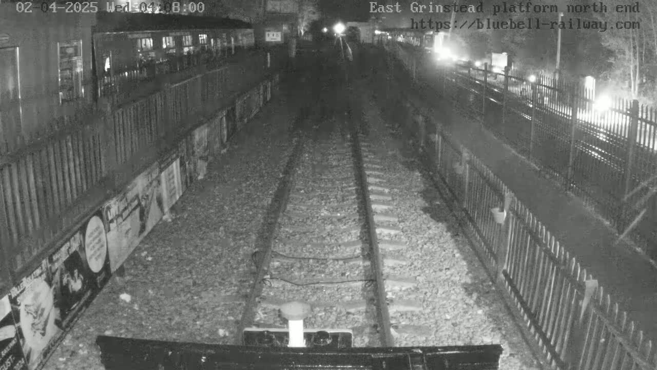 A nighttime view of train tracks at a station platform, bordered by fences and posters, under dark conditions.