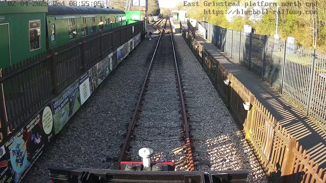 A view from a low vantage point down a railway track on a sunny day, showing a green train car and wooden fences on either side.