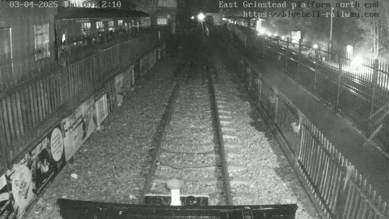 A nighttime view of train tracks at a station platform, with fences and posters visible along the sides.