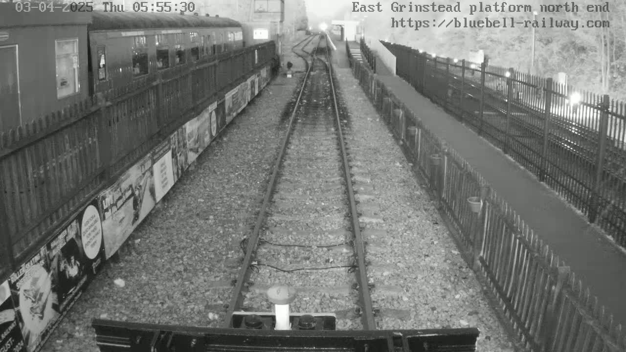 A nighttime, grayscale view from a train shows a railway platform with a train car to the left and fences on both sides of the tracks.