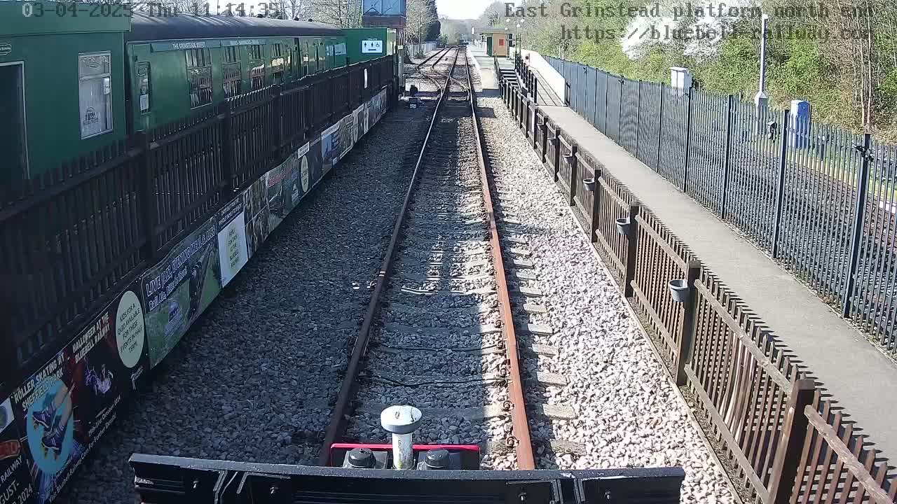 A view from a train's perspective down a sunny railway track, flanked by fences and posters, with a green train car visible to the left.