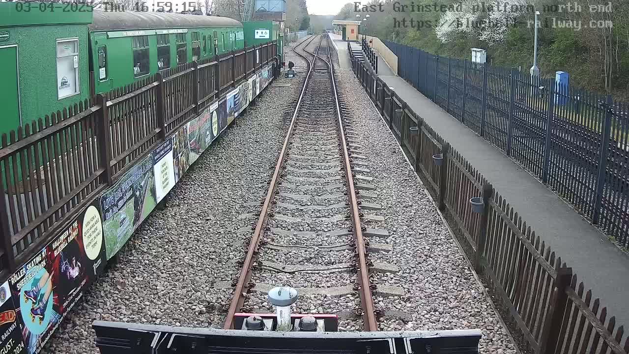 A view from a train's perspective shows railway tracks extending into the distance, flanked by a green train car and wooden fences on either side, under a partly sunny sky.