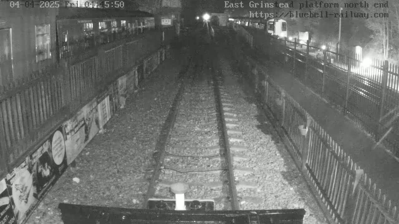 A nighttime view down a set of train tracks, bordered by fences and posters, at a seemingly empty station platform.