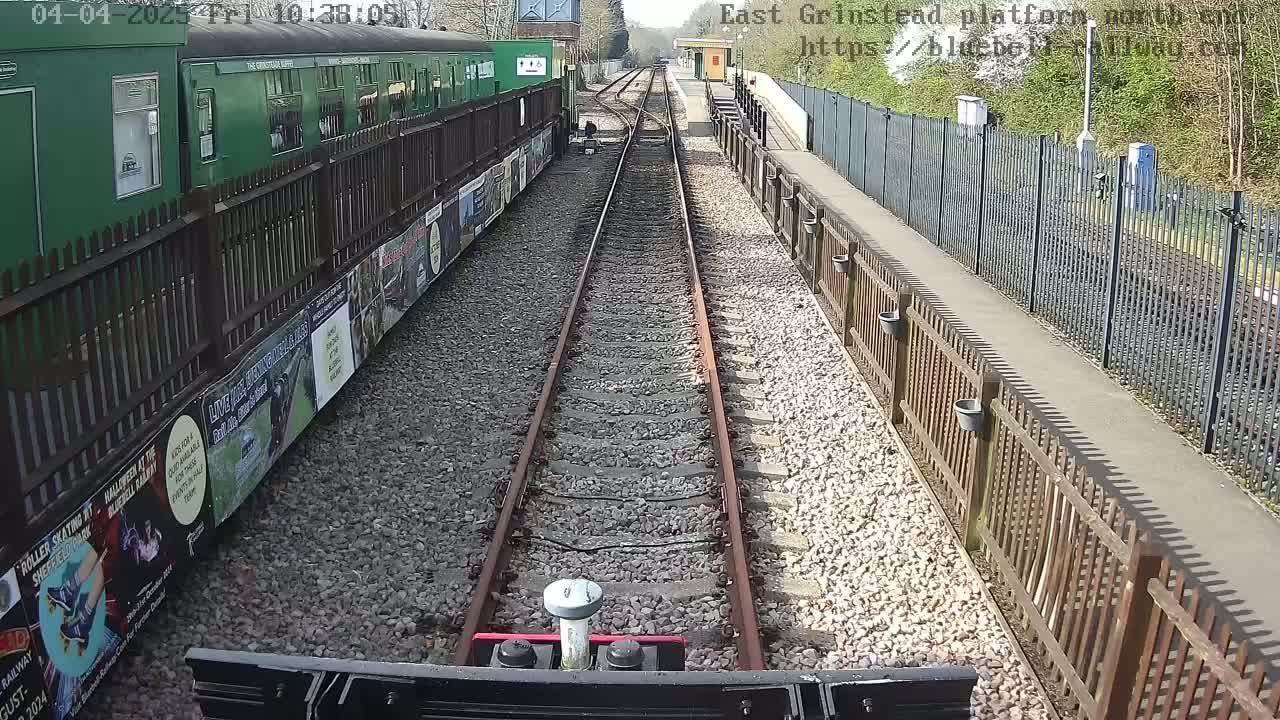A view from a train's cab shows a sunny day at a railway station with a green train car and posters on a fence alongside the tracks.