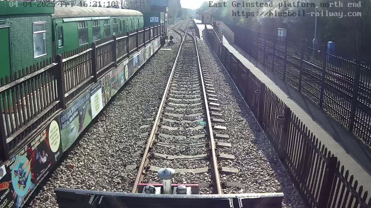 A view from a train shows a sunny day, railway tracks extending into the distance between fences lining a station platform, with a green train car alongside.