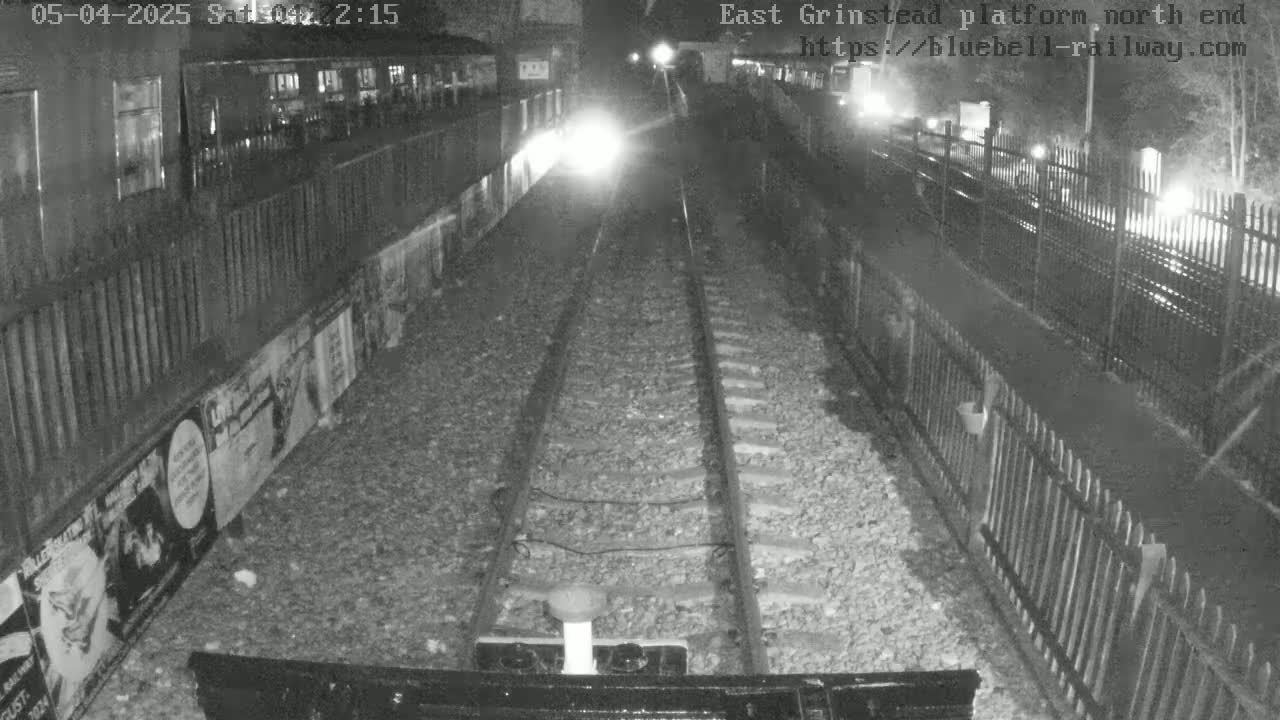 A nighttime view of train tracks at a platform, bordered by fences and posters, under dark conditions.