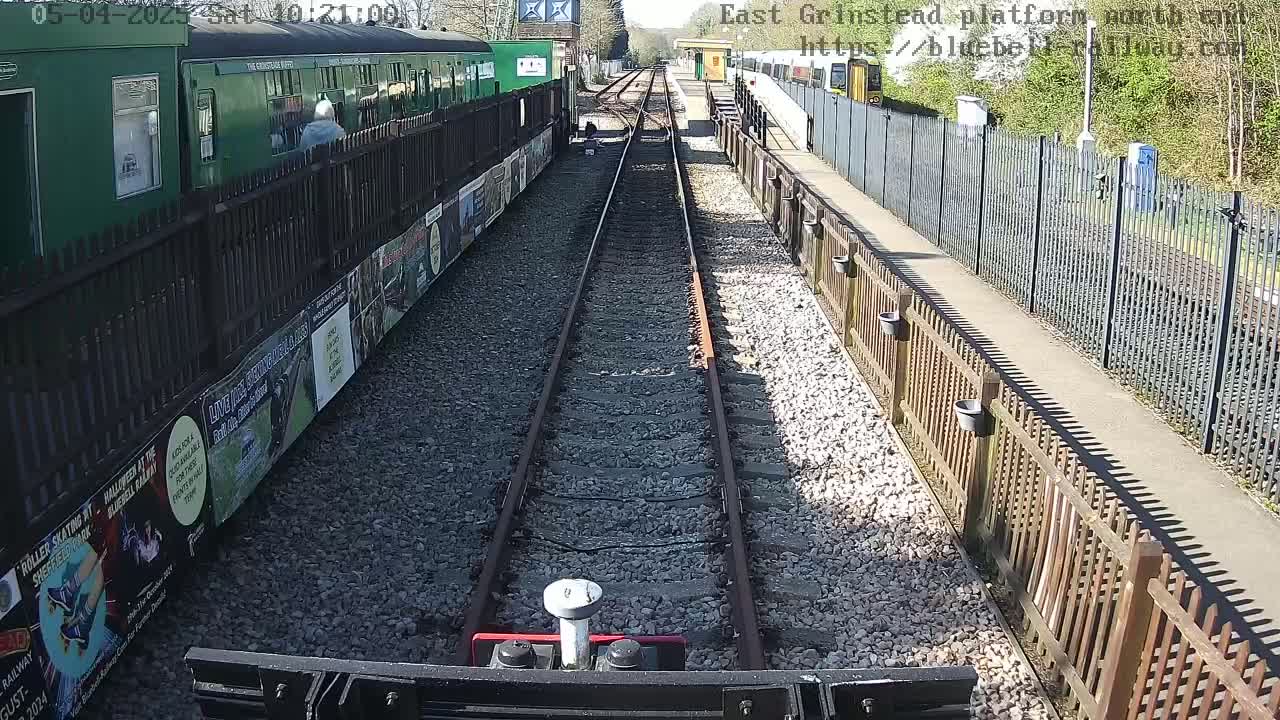 A sunny day at a train station with a view down a railway track between two platforms, one with a green train and the other with a yellow train, bordered by fences and posters.