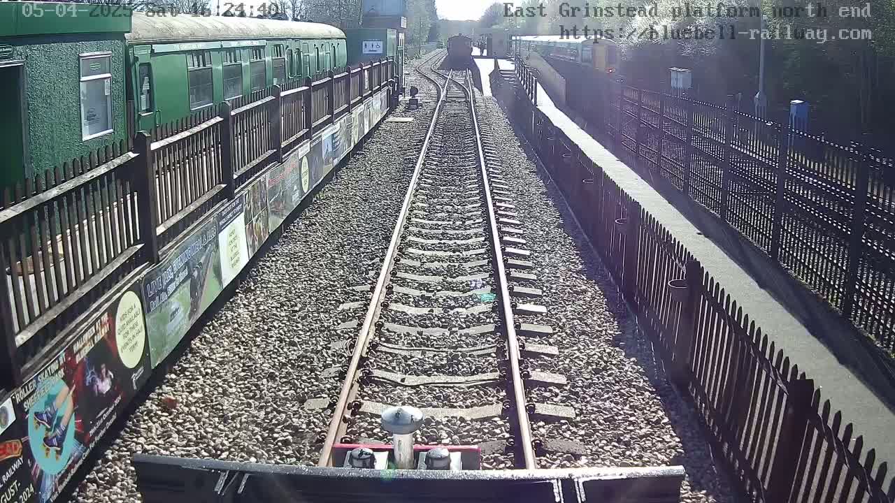 A sunny day at a train station shows a view down a railway track between fences, with a green train car and another train visible in the distance.