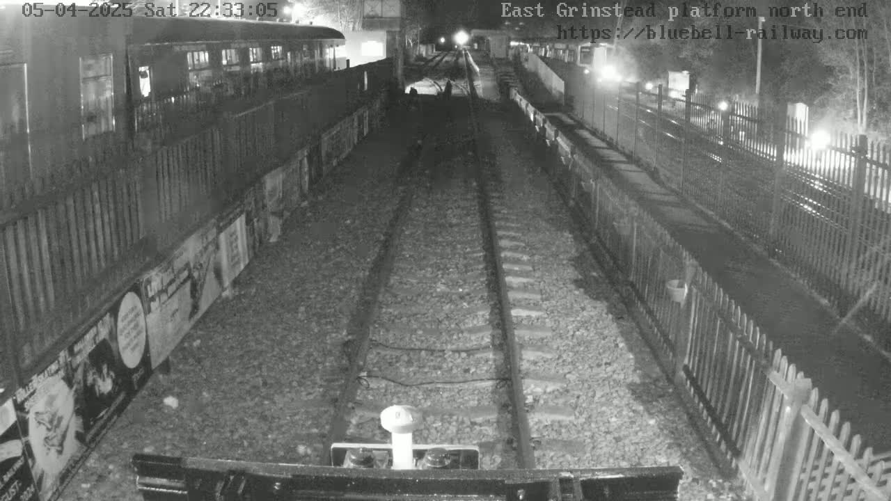A nighttime view from a train shows a dark platform with railway tracks extending into the distance, alongside fences and posters on the walls.