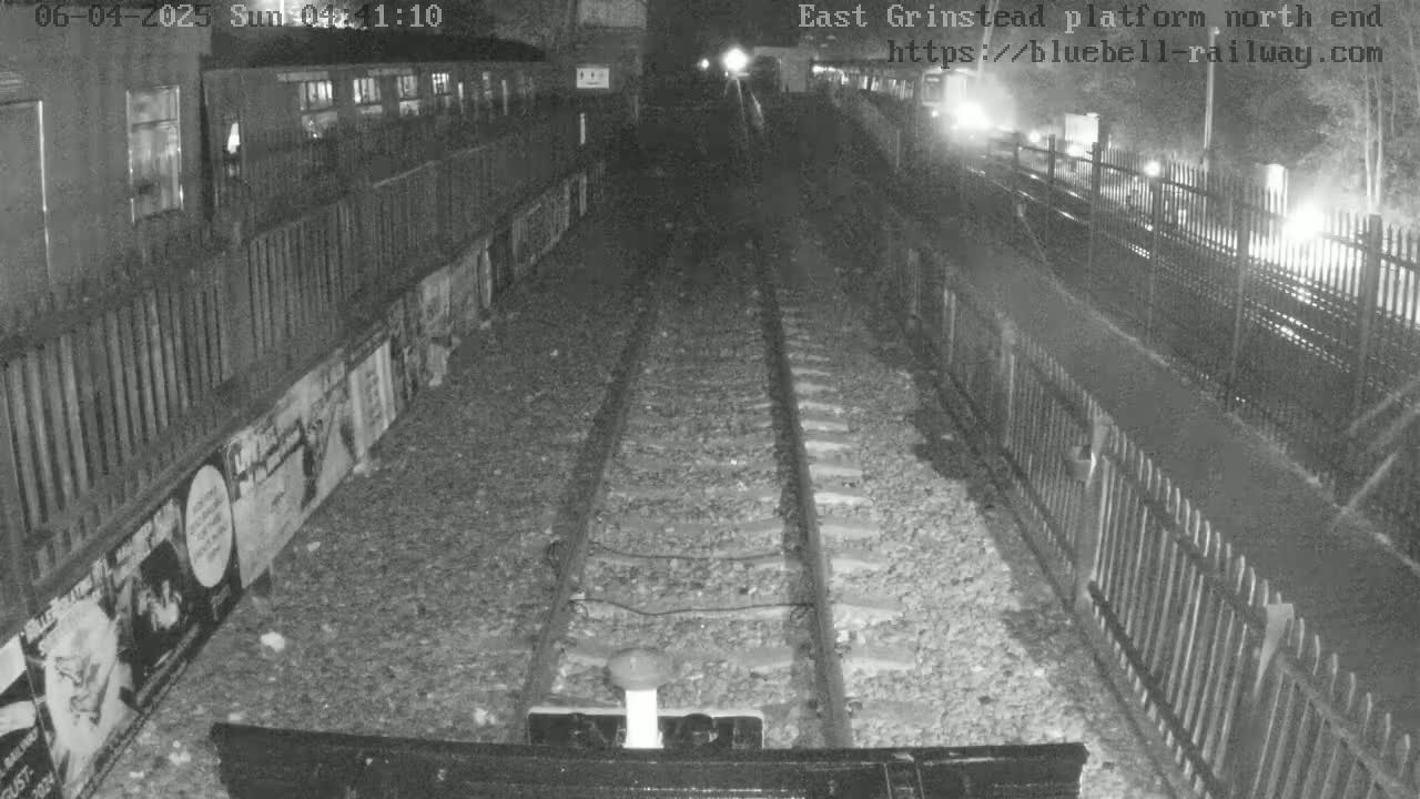 A nighttime view of train tracks at a station platform, with posters on the wall and fences on either side.