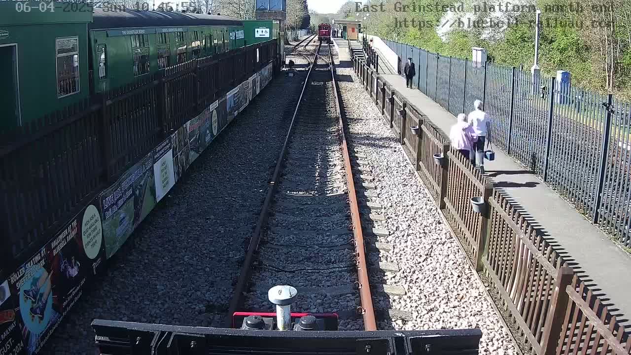 A sunny day at a train station shows a red train in the distance, two people walking on the platform, and a green train alongside the platform.