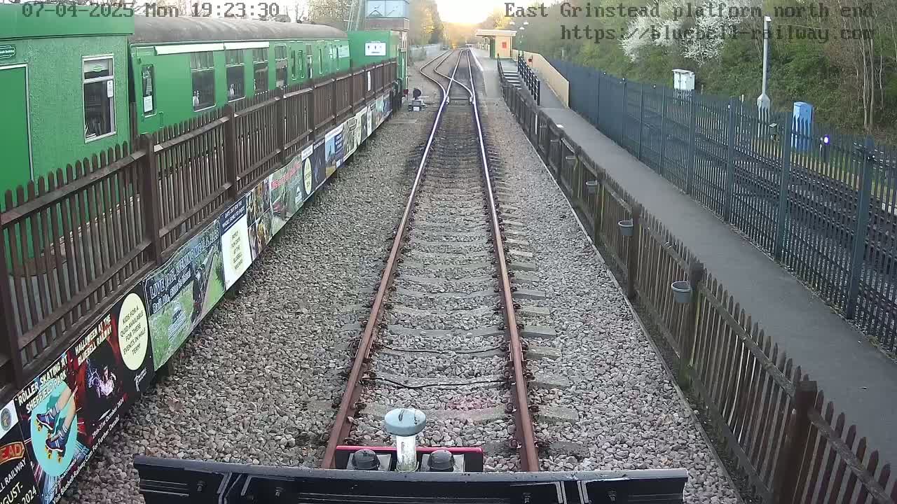 A view from a train's cab shows two sets of train tracks leading to a station platform under a twilight sky.