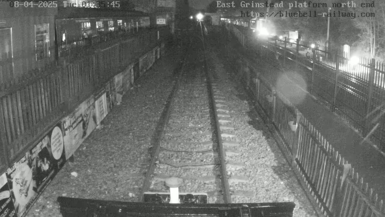 A nighttime view shows train tracks extending into the distance between fences lined with posters, under dark, possibly cloudy skies.