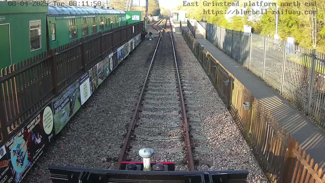 A railway track stretches toward a sunny, green landscape, flanked by wooden fences and a parked, green passenger train with posters along its side.