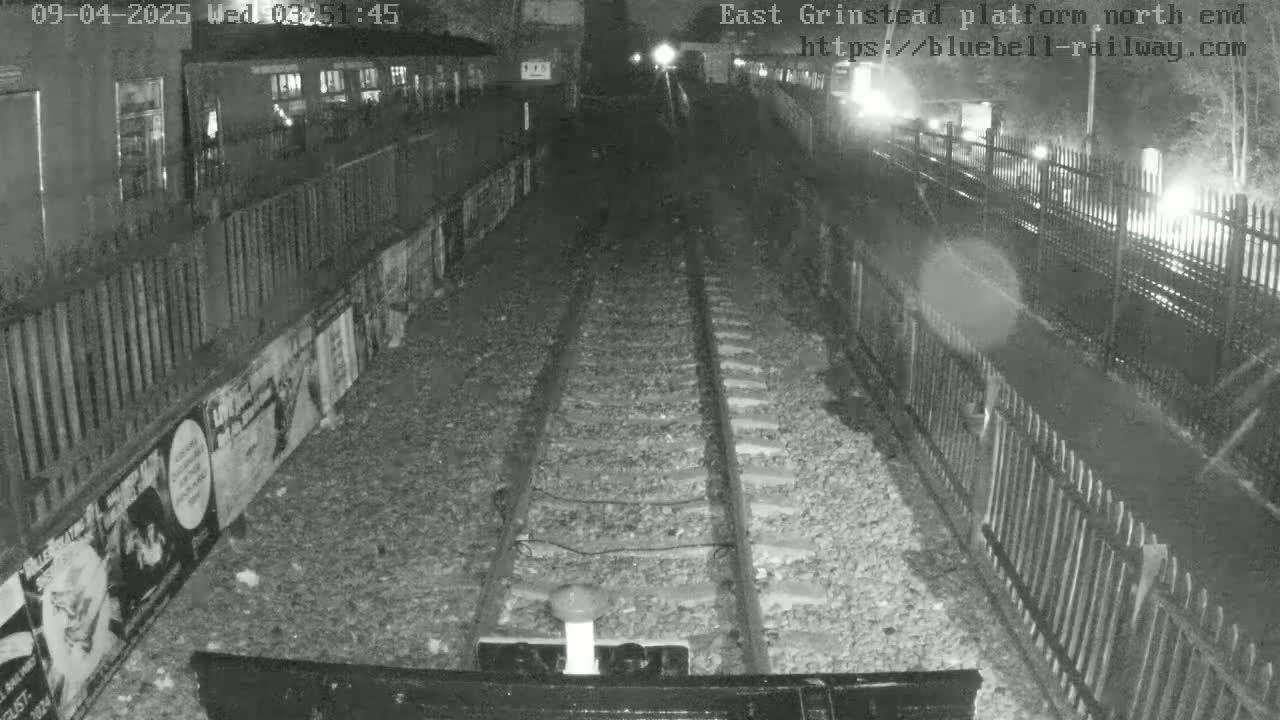 A nighttime view from a train shows a railway platform with fences, posters on the wall, and train tracks extending into the distance.