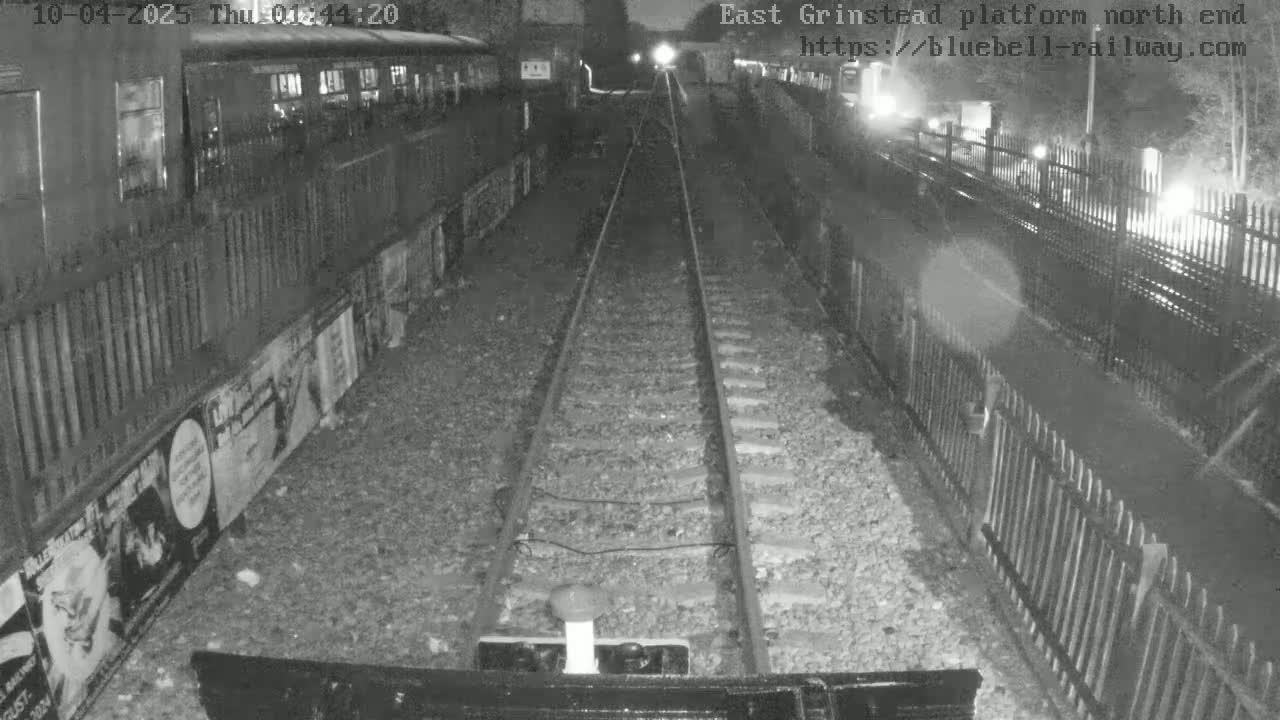 A nighttime view of train tracks extending into the distance, flanked by fences and a train car to the left.