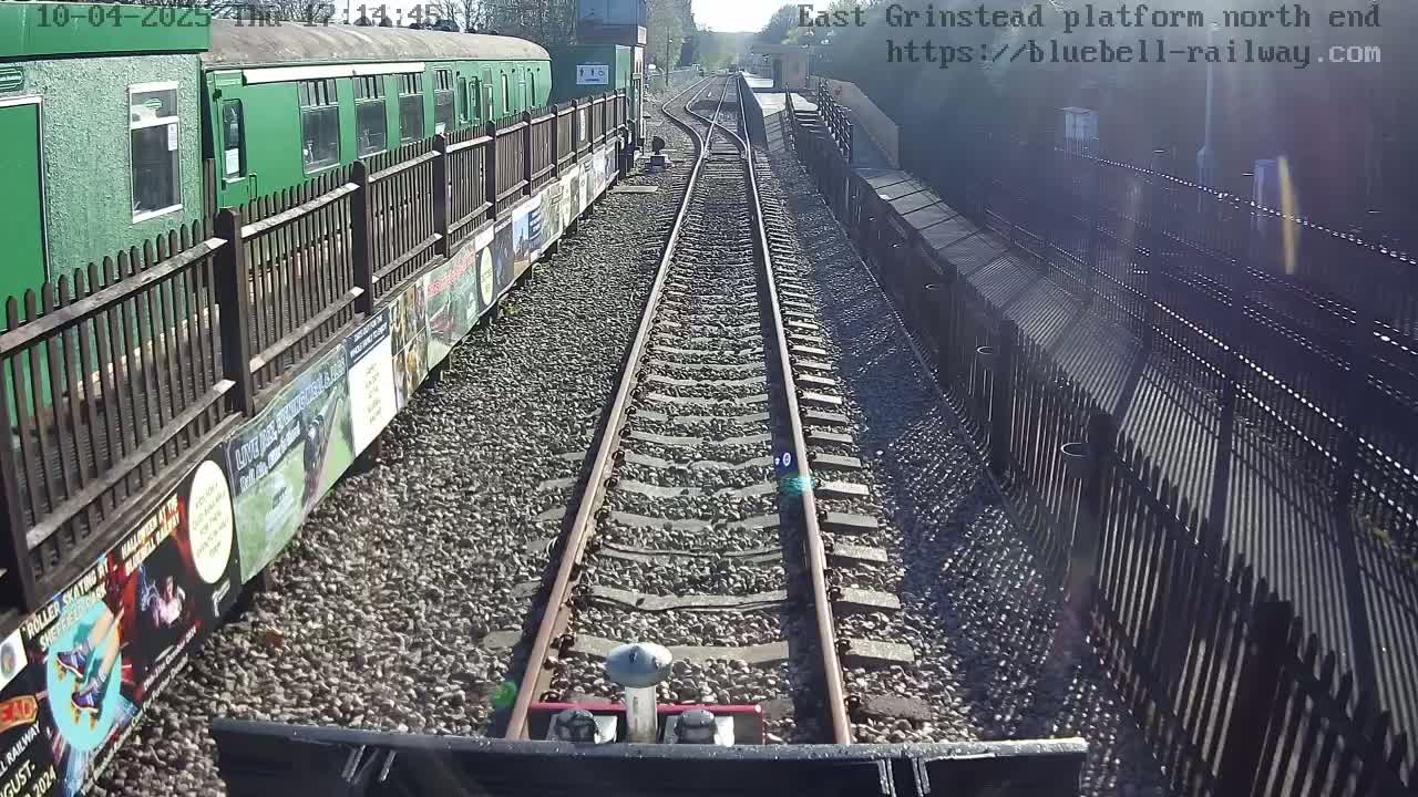 A train travels down a railway track on a sunny day, alongside a platform with a green train and wooden fences.