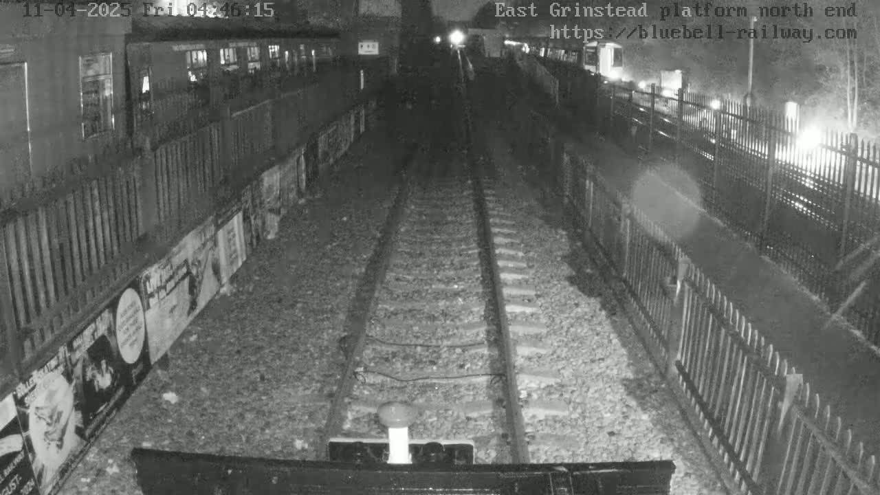 A nighttime view of train tracks at a station platform, with fences and advertisements lining the sides.