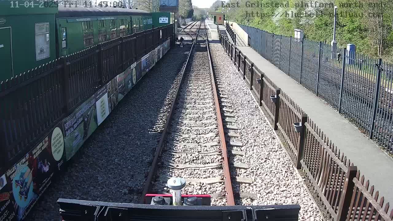 A view from a train's cab shows a sunny day at a train station with railway tracks stretching into the distance, flanked by fences and posters on the side of a green train.