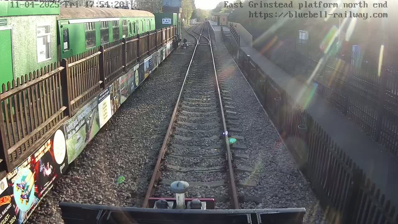 A view from a train's cab shows two parallel train tracks extending into the distance on a sunny day, with a green train car and a wooden fence on the left, and another fence along the right side.