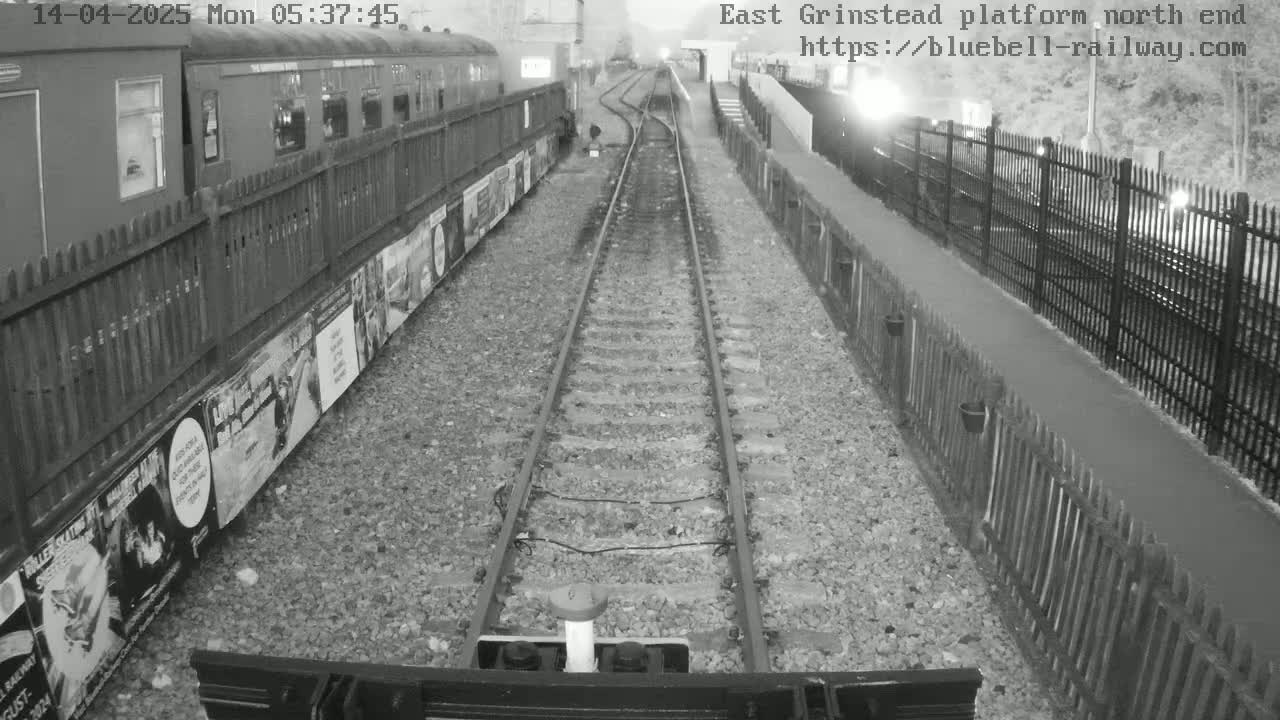 A nighttime view of railway tracks extending from a train car parked alongside a platform, bordered by fences and posters.