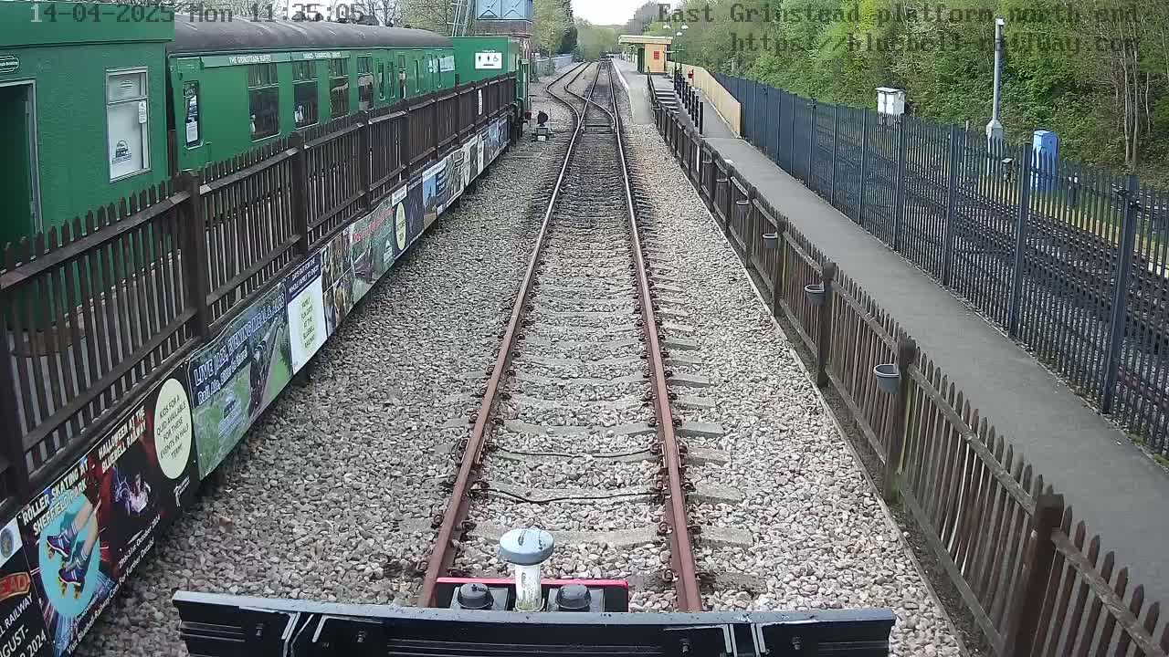 A train travels down a railway track on a sunny day, passing a green train car and a station platform with fences and posters.