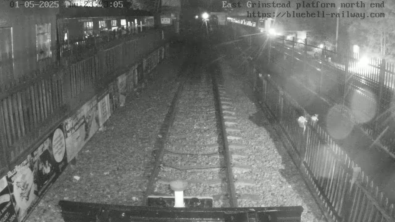 A nighttime, low-light view of train tracks at a platform, with fences and posters lining the sides.