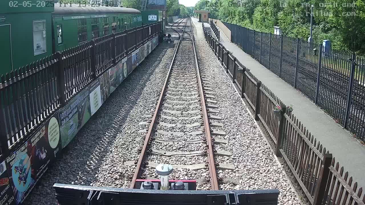 A train travels along a railway track on a sunny day, passing alongside a platform with a green train and wooden fences.