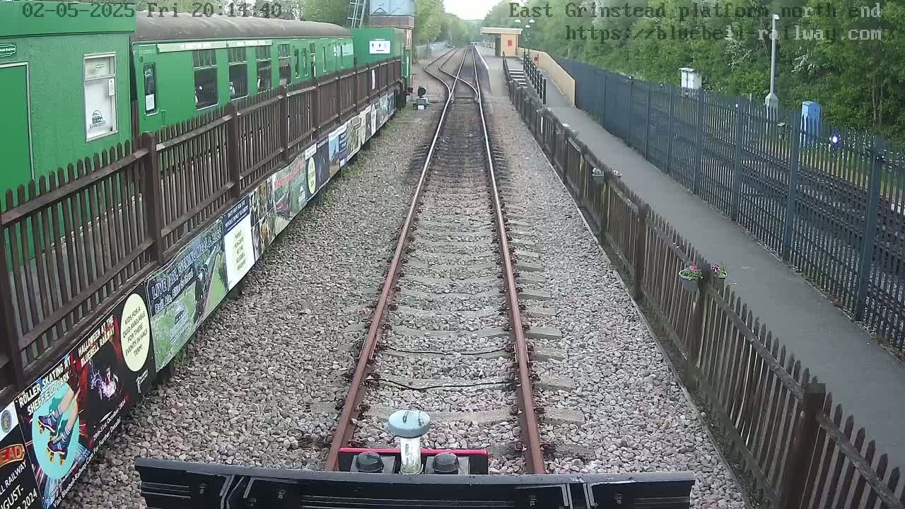 A view from a train shows a railway track extending into the distance, flanked by wooden fences and posters, with a green train car visible to the left.