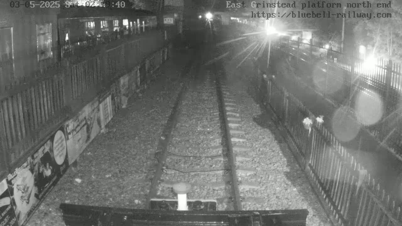 A nighttime view of train tracks extending into the distance between fences, bordered by posters on one side.