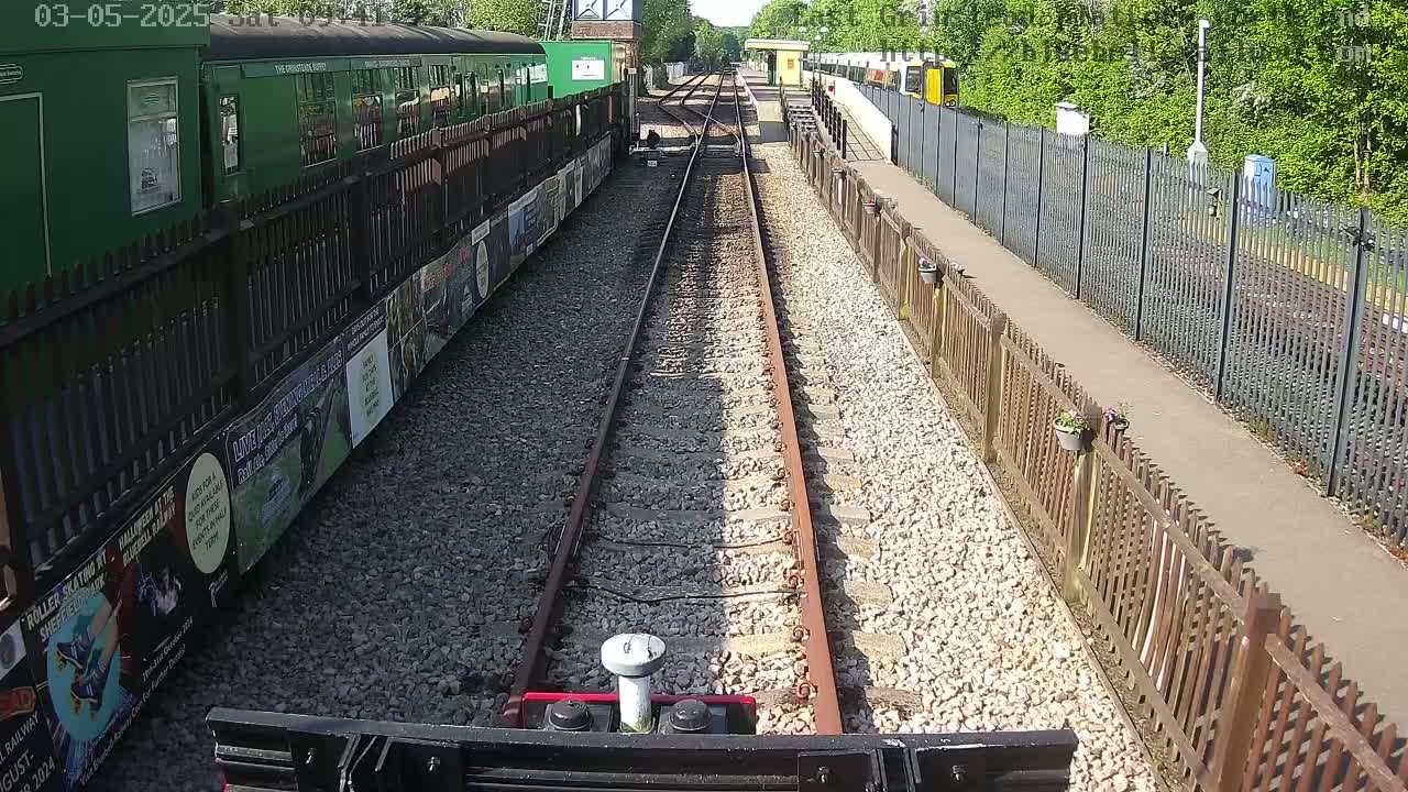 A view from a train's perspective shows railway tracks on a sunny day, alongside a green train and wooden fences, with a gravel path and additional trains visible in the distance.