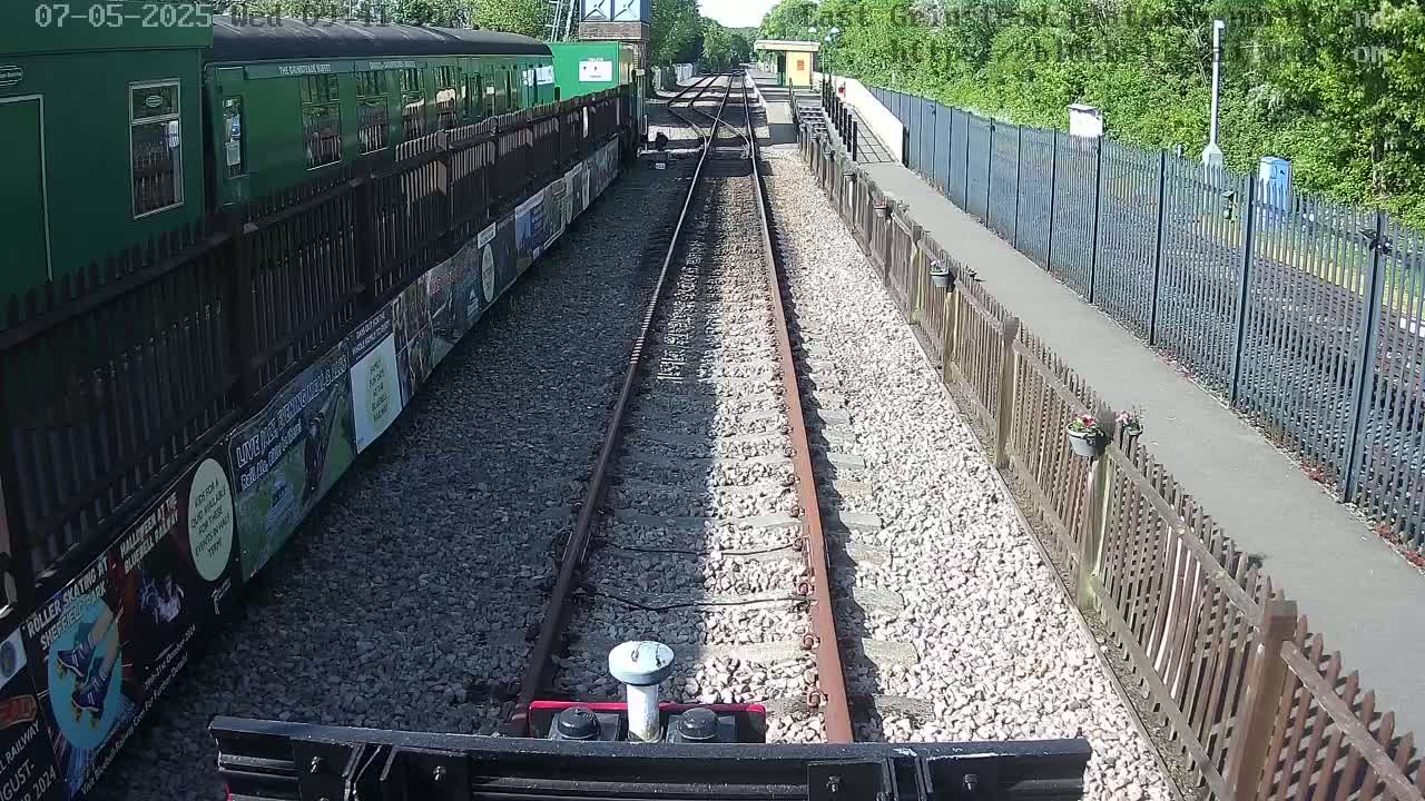 A view from a train shows a sunny day at a railway station with a green train and wooden fences alongside the tracks.