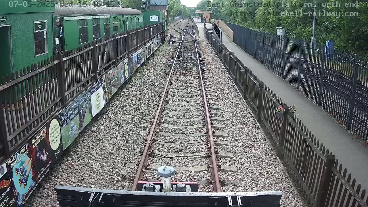 A view from a train's perspective shows a railway track extending into the distance, flanked by a green passenger car and wooden fences, on an overcast day.