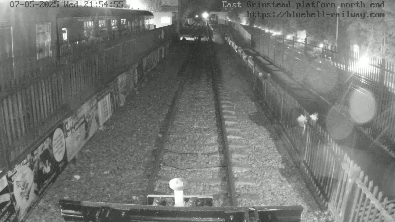 A nighttime view of train tracks at a platform, bordered by fences and posters, with a train car partially visible in the background.