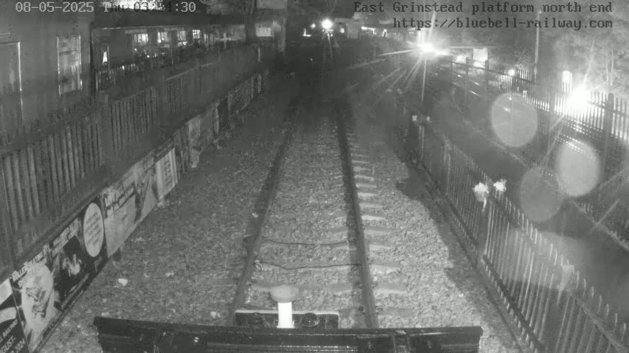 A nighttime, low-light view from a train shows a railway track extending into the distance between fences, with posters visible on a wall to the left.