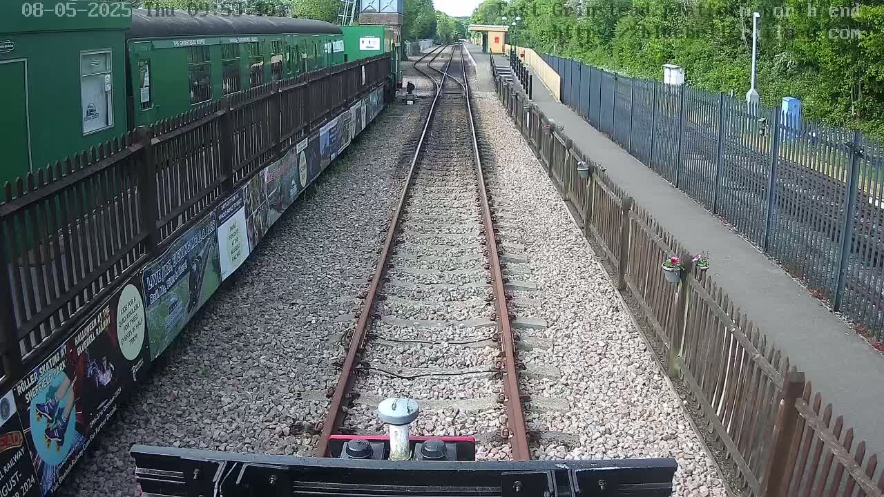 A railway track extends from a green train, bordered by fences and lush greenery under a sunny sky.