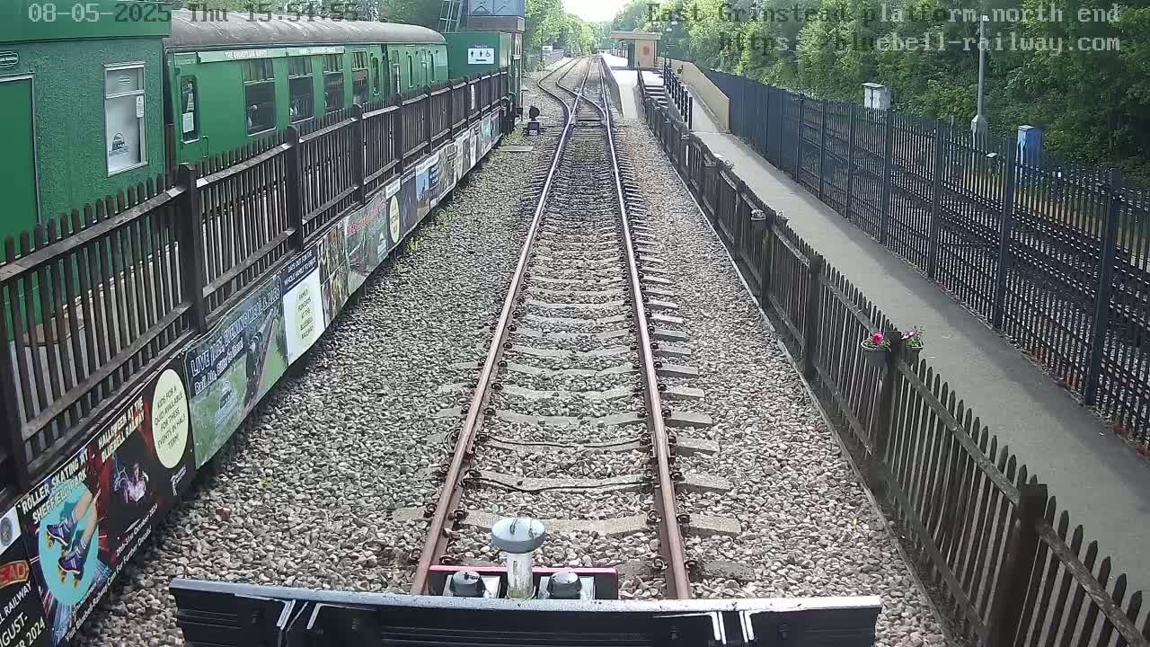 A train track lined with a green train car and wooden fences stretches into the distance on a sunny day.