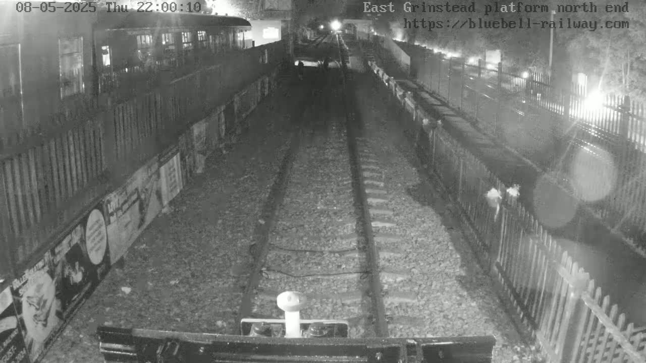 A nighttime view from a train shows a railway track extending into a tunnel, flanked by fences and posters on the platform walls.