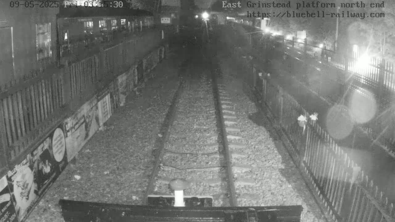 A nighttime, low-light view of train tracks extending into a tunnel, flanked by fences and walls with posters.