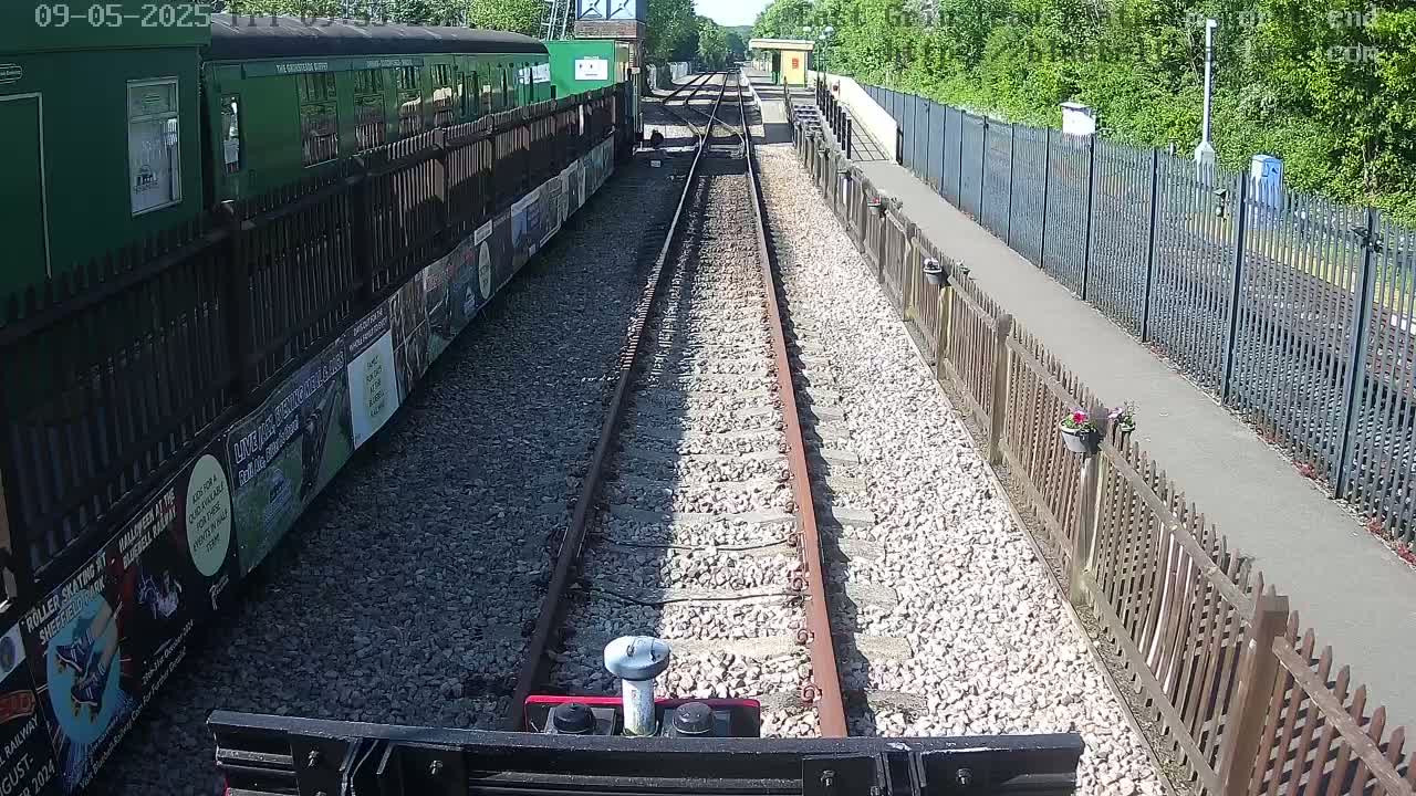 A railway track extends from a train and platform, bordered by fences and gravel, under a sunny sky.